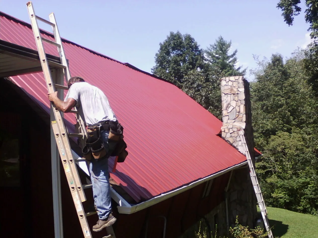 Roofer on a ladder beside a red metal roof and stone chimney — Soffit & Fascia Repair by Roofing Brothers in Lenox